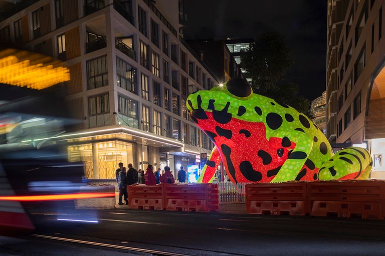 A giant inflatable neon green and red frog sculpture illuminated at night on a city street next to modern buildings and moving traffic.
